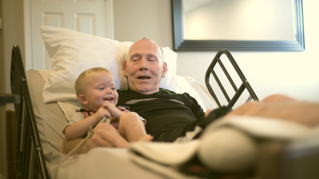 photo of older man in hospital bed at home with young child sitting next to him